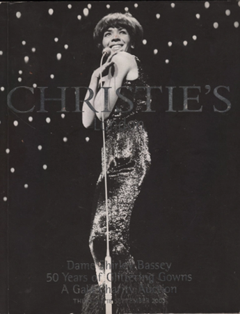 A reproduction of a poster for a gala charity auction by Christie’s auction house, featuring a black and white photo of singer Dame Shirley Bassey, dressed in a sparkling dress and standing by a microphone. The text on the poster reads: “Christie’s. Dame Shirley Bassey. 50 years of Glittering Gowns. A Gala Charity Event Auction. Thursday 18th September 2003.”