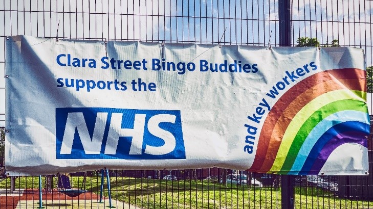 A photograph of a large banner hanging on a railing, with an image of a rainbow and the NHS logo (large white letters inside a blue rectangle). The text on the banner says: “Clara Street Bingo Buddies supports the NHS and key workers.”
