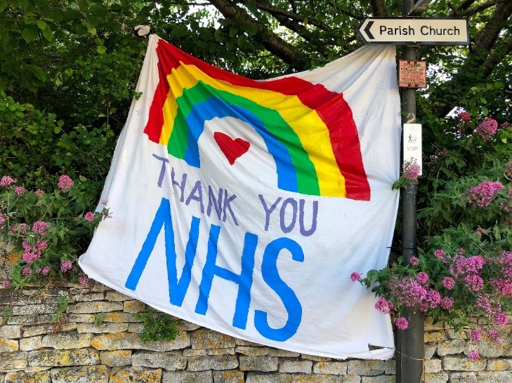 A photograph of a large textile banner hanging between a tree and a lamp post. The banner features the image of a rainbow and a heart, with the letters: “Thank you NHS.”