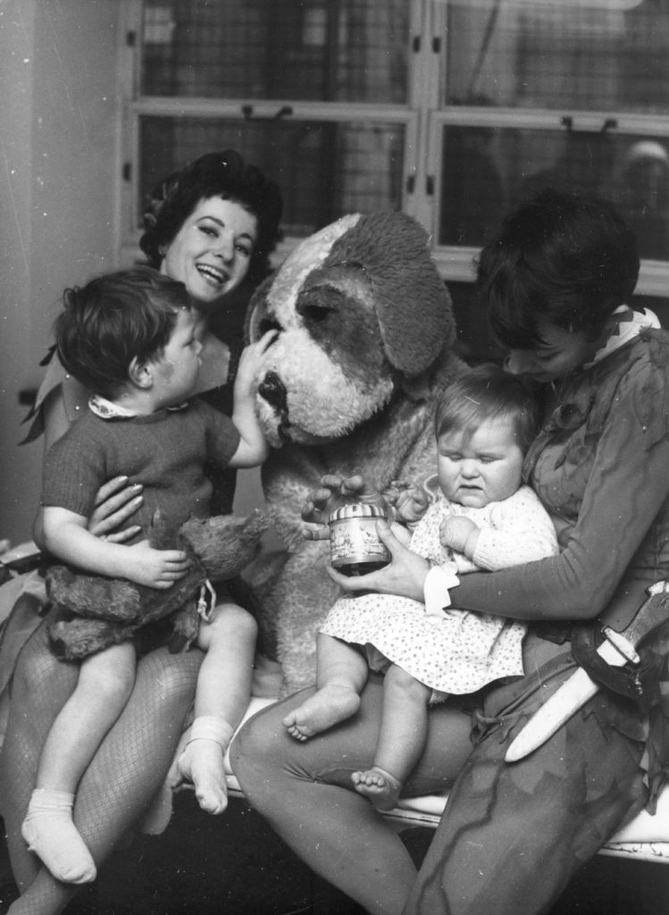 Photograph of the cast of Peter Pan visiting patients at Great Ormond Street Hospital. There are three cast members in the photo - Tinkerbell, Peter Pan, and Nana the dog. All three are in costume. There a two children, one sat on Tinkerbell's lap whilst stroking Nana's face, and one sat on Peter Pan's lap.