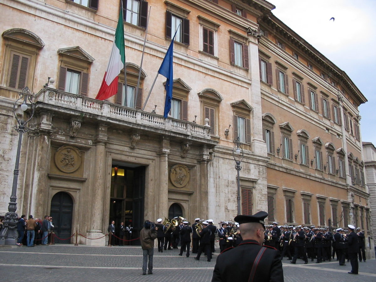 The front entrance to the Italian Parliament. It is a three floored pink stucco building of 18th Century type vintage. Slightly grubby and hard to decipher carvings line the doorway in a dark stone. The Italian tricolor and the flag of the European Union hang limply from flagpoles over the doorway. On the square outside a military or police band are filing in. They hold brass musical instruments and are dressed in very dark blue/black uninforms and hats. Their uniform has a thin red trim to it