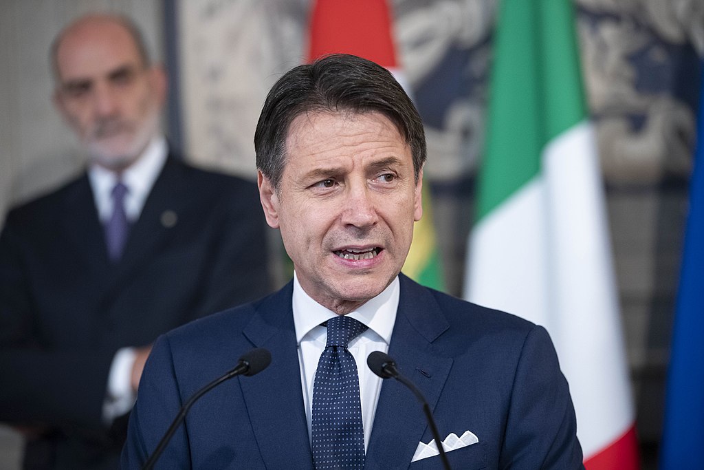 Giuseppe Conti (5 Star Movement affiliated Prime Minister of Italy since 2018) stands at a lectern midst giving a speech. He is a white male of around fifty years of age, has greying dark hair and a slightly tanned face. He is clean shaven and isn't wearing glasses. He wears a dark blue suit with a white handkerchief in to top pocket. His tie is a similar dark blue colour and his shirt is white. Behind him a figure of a male aide or another politican is visible, he is also wearing a suit, aged between 50 and 60, but is balding with a beard. the wall behind both men is marble and an Italian tricolor is partly visible on a flagpole