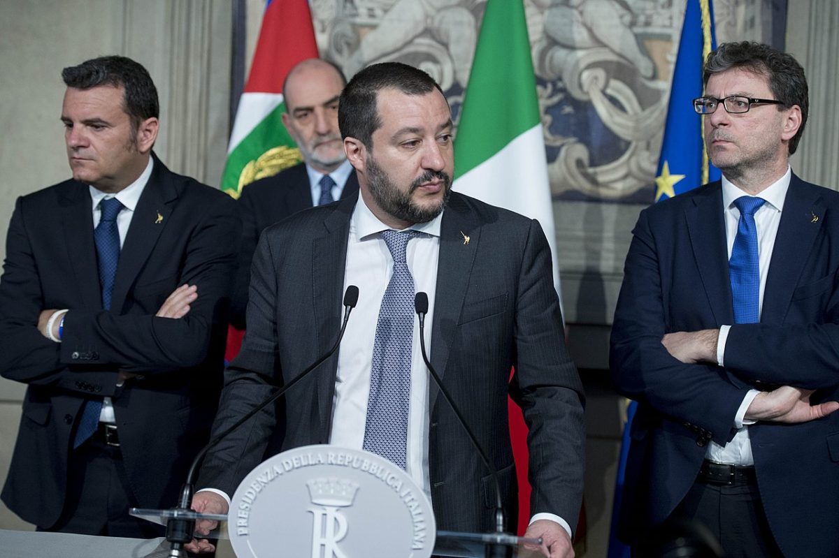 Matteo Salvini stands at an official Italian government lectern giving a speech. He wears a dark grey suit and lighter grey tie, with a white shirt. He is flanked by two men who are similarly attired with another male figure stood behind them. part of a stone carved statue can be seen behind them alongside several Italian tricolors and an EU flag