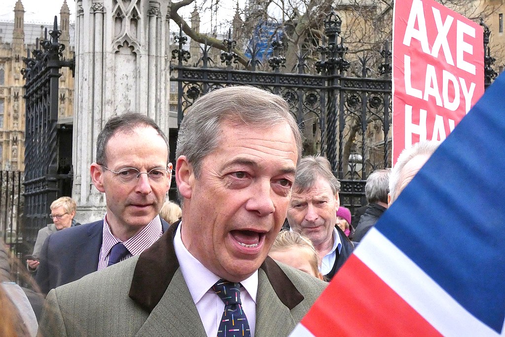 Nigel Fararge depicted outside the gates of the Houses of Parliament in January 2019. He wears a green overcoat with a brown collar as well as a pink shirt and a dark blue tie with white dots. Part of a unon jack is in the foreground of the shot, a protest placard can be seen behind him and there is a cluster of midde aged to elderly people, mostly male stood behind him. Nigel Farage has his mouth open as if he is talking to someone who is out of shot