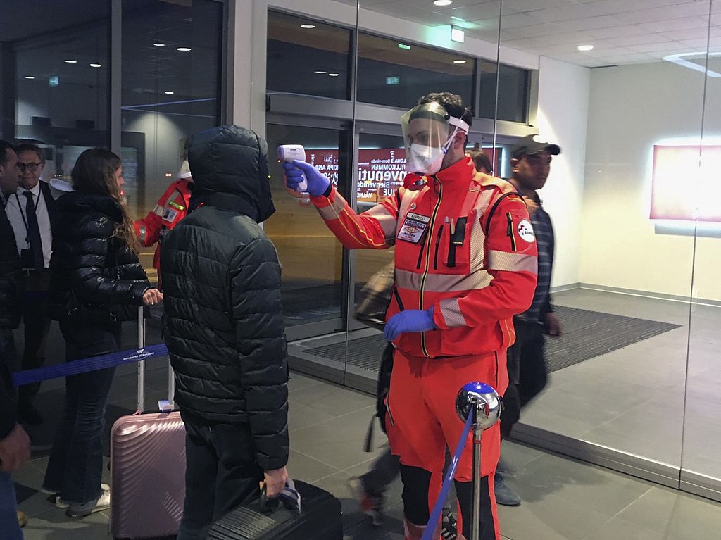 Passengers have disembarked plane and are moving through an airport termina building. A youn white man aged about 30 in a fluorescent orange suit wearing boots, gloves, a visor and facemask is stood taking people's temperatures as a Covid-19 screening and safety precaution