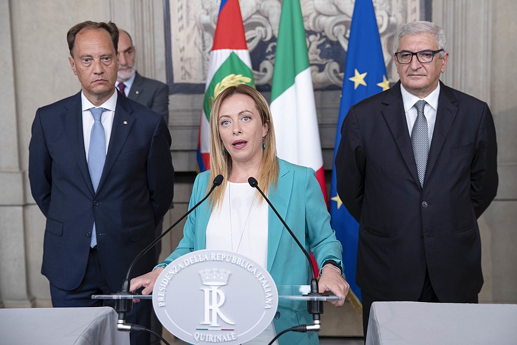 Giorgia Meloni (a white woman in her early 40s) stands in front of a podium. She is wearing a teal coloured jacket and a white blouse. She is flanked by two white middle aged men probably in their 50s both of whom wear suits and ties. An Italian flag and an EU flag hang upright on poles behind them