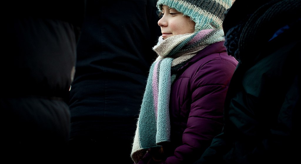 Greta Thunberg is pictured in profile stood amongst a group of people in back coats. She wears a dark purple winter coat and a coat and a hat which are green/blue, white and purple striped in colour