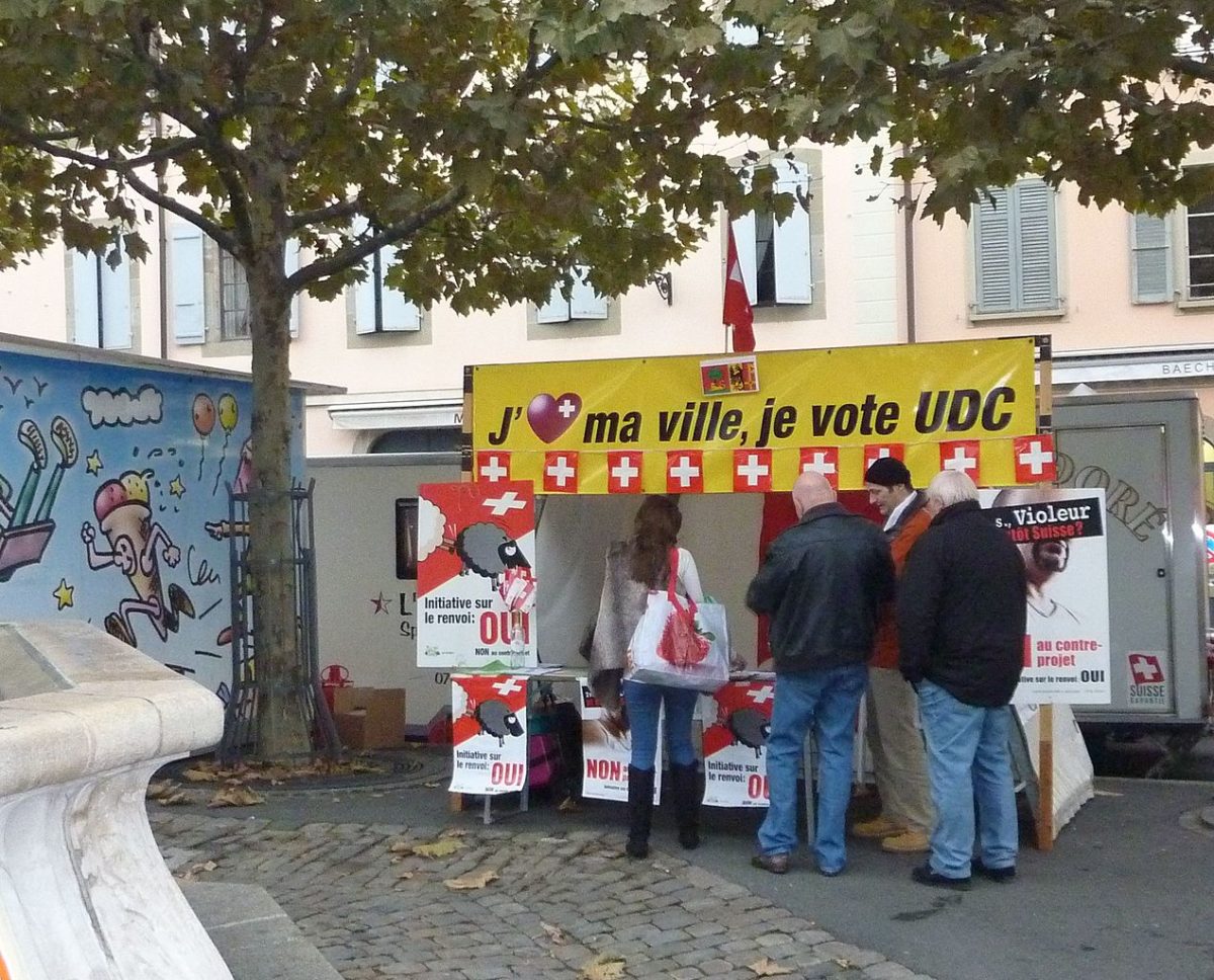 Street stand for the Swiss people's Party. A group of middle aged white people, affluent looking, all men apart from one woman, stand with their backs to the camera deep in discussion. They stand at the side of a small town square under some trees on an early autumn day, in front of a market stall style stand bearing Swiss flags and Swiss People's Party posters