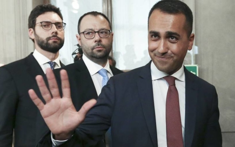 The Five Star Movement’s former leader Luigi Di Maio. Depicted wearing a suit and tie and standing. Two men with glasses also in suits stands behind him