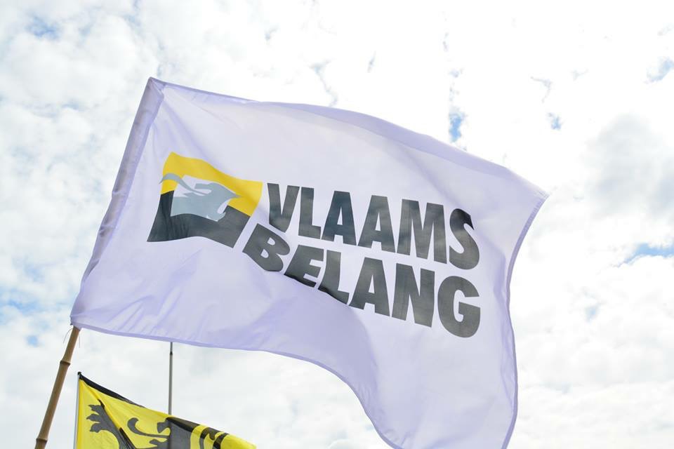 Vlaams Belang logo on a white flag fluttering aloft on a cloudy day. The flag bears both the party's full Flemish name in black block capitals alongside its logo, a black and white crest imposed on the Flemish flag (black and yellow). A small portion of a Flemish regional flag can be glimpsed in the lower part of the frame