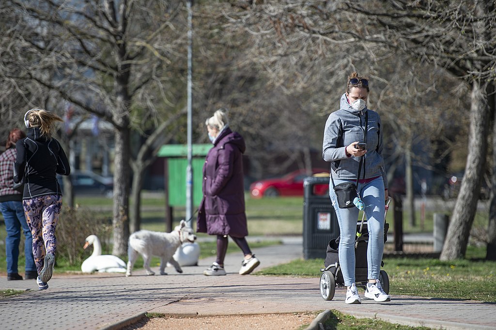 Image is of a park in Bratislava capital of Slovakia. It is a cold but sunny spring day. Four white women are pictured walking along paths crossing the park which is lightly wooded and grassed. Its near a lake an there is a small nuber of geese on one of the paths. A large green wheelie bin sits in the middle of the image. All of the women, who are different ages, are walking separately and not connected with each other. They're wearing facemasks in compliance with Covid-19 public health restrictions