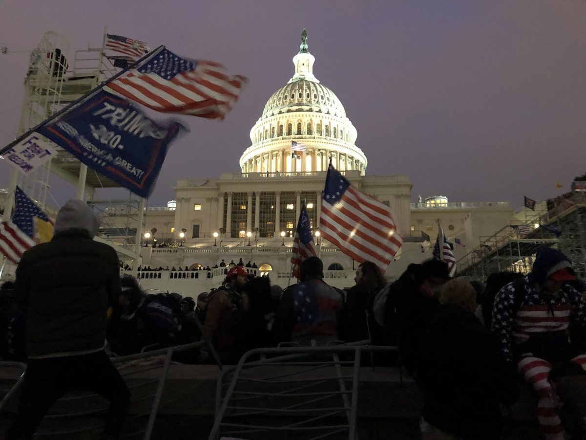 US Capitol Building at dusk. The dome of the building is illuminated in the centre of the image. In the foreground protestors in winter clothing many waving Trump flags, and flags representing the early flag of the United States c. 1785 are flying. A large crowd is assembled stretching up to building which is barricaded with crowd control barriers. A group can be seen on the building's terrace, it's not clear if these are soldiers/police or protestors