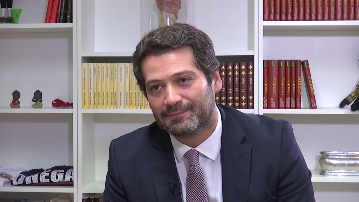 Picture of Chenga (Enough!) party leader André Ventura. He is a white man in his late 30s with greying mid-coloured brown hair, a short greying beard of the same complexion and is seated in front of a bookcase which contains several rows of books organised by series and some knickknack type items including a folded Chenga t-shirt. He is wearing a navy/black coloured suit jacket, white buttoned up shirt and a salmon coloured tie