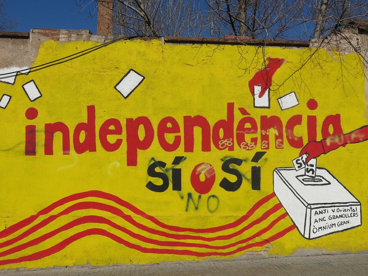Outdoor mural ahead of the Catalan Independence Referendum in 2017. It has the words "Impendence Yes, Yes" written in red and black on a yellow painted wall. The image also includes disembodied red hands placing white ballot papers into a white ballot box which has Catalan text written on it. The mural has been slightly defaced by someone crossing out the Catalan "Si" for "Yes" and writing "No" in English in black spray paint