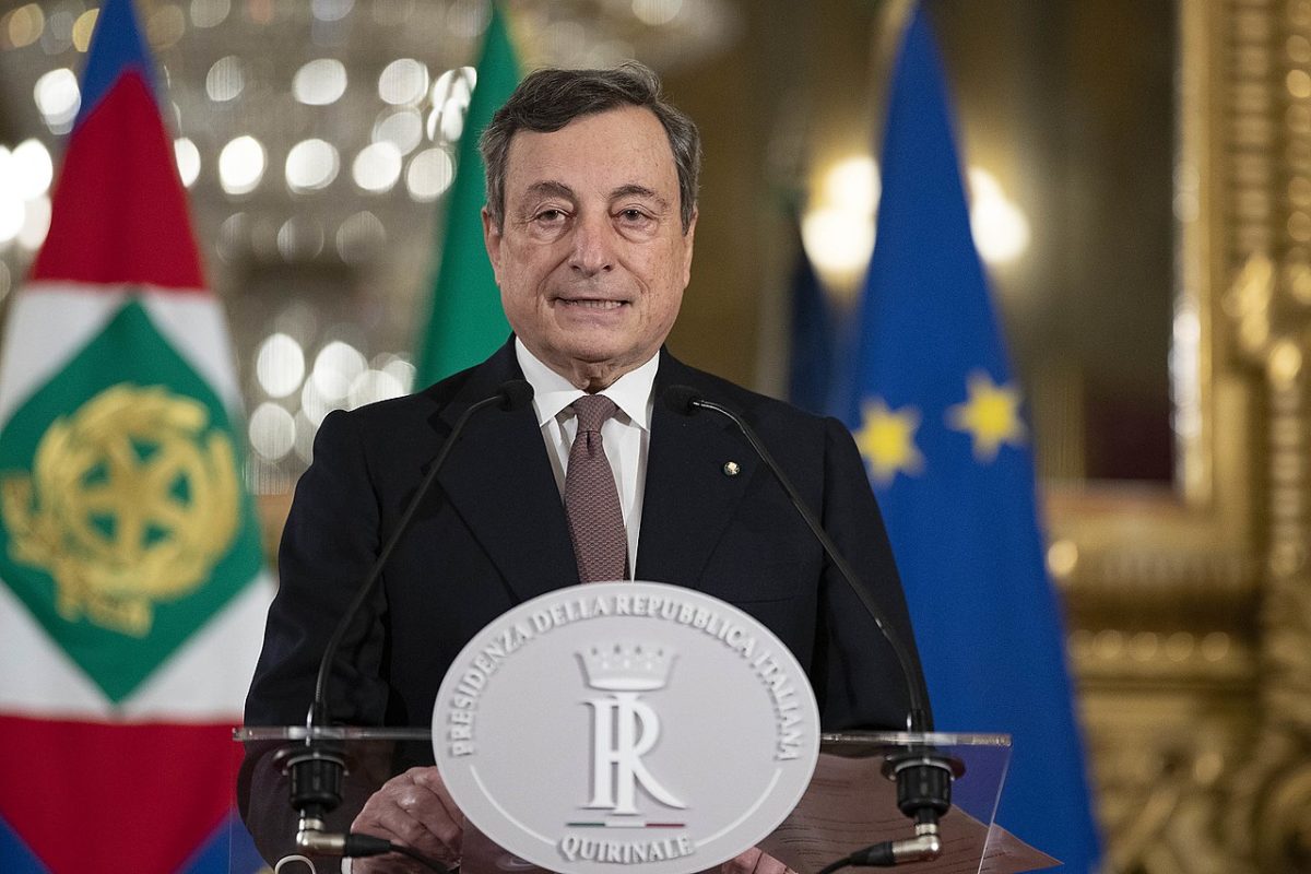 Italian premier designate Mario Draghi stands at a podium in the Quirinale Palace. he is an elderly white man in his 70s with short dark hair, wearing a black suit, white shirt and a salmon coloured tie with a dark spotted pattern. His podium has a silver logo of the Italian government embossed on the front. Behind him chandeliers, an Italian tricolour and an EU flag can be glimpsed