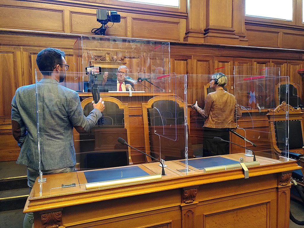 An elderly white man in a suit and tie sat on a diasis in a wood panelled hall is interviewed by two white journalists, one is man, one is a woman, both of whom are also smartly dressed and who are in their late 30s. Bundeshaus (Bern / Switzerland) with plexiglass protection against Covid-19: President of the Council of States Hans Stöckli in an interview in the Chamber of States