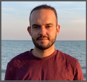 Profile picture of Mattia Zulianello. He is a white man in his 30s with dark hair pulled back and a short full, dark beard. He is stood on a beach at twilight with the sea behind him and wears a claret coloured t-shirt.