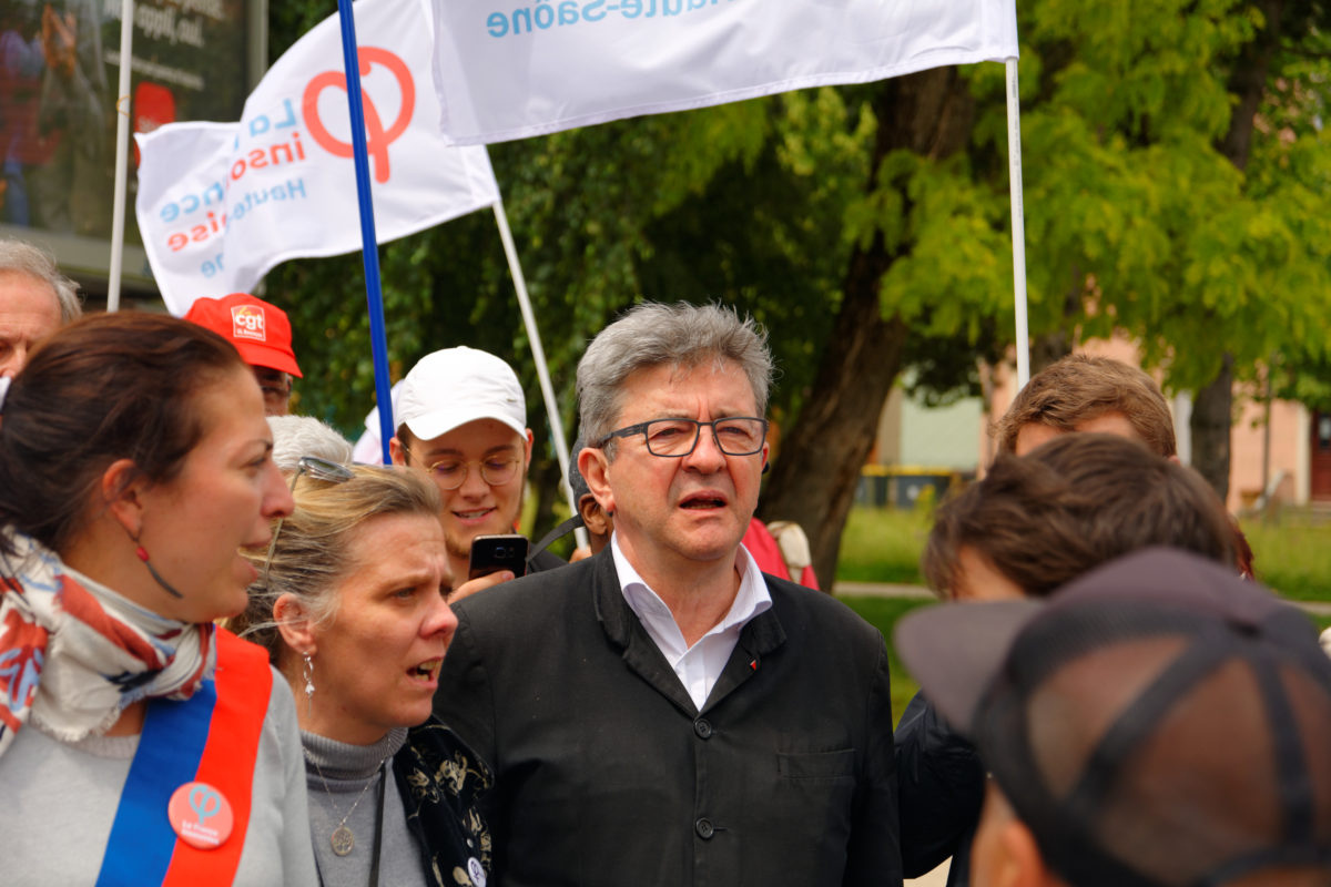 Jean-Luc Mélenchon is depicted amidst a group of striking workers. The striking workers are a range of ages, mostly younger white women, and are stood outside on a sunny day holding flags representing their trade union as if picketing or at a rally. Jean-Luc Mélenchon is stood the centre of the frame. He is a white man in late middle age with largely white and grey hair, clean shaven with glasses. He wears an open neck white shirt and a largely buttoned up workers' style jacket in black with a couple of small pin badges on the lapel